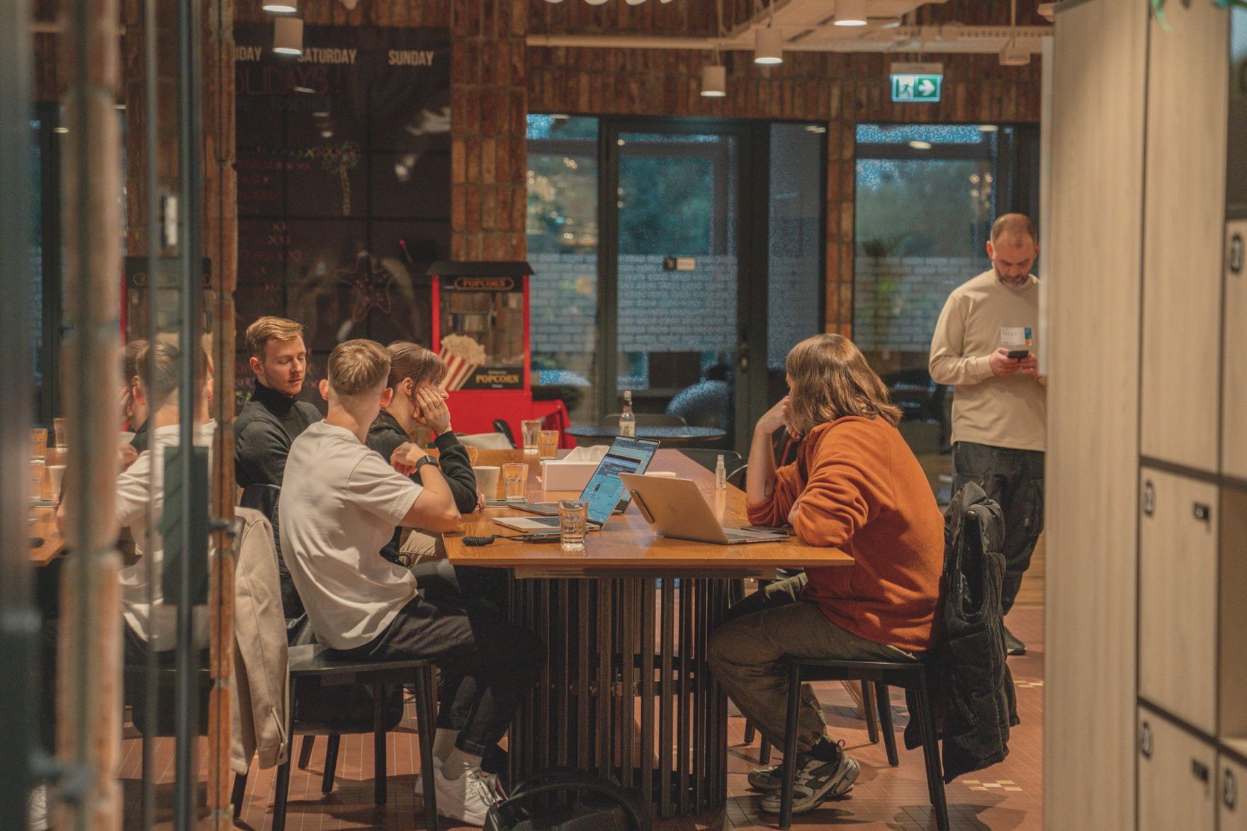 A long table in a brick-walled room, six people working on laptops, one standing beside them.